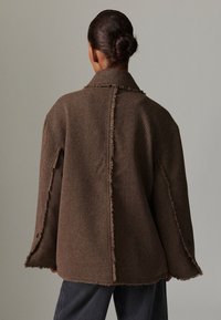 Brown oversized jacket with a textured finish, frayed edges, and a collar. Features wide sleeves and button details at the cuffs.
