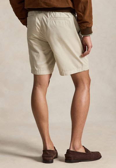 Moški v bež kratkih hlačah, rjavih semiš loaferjih in rjavi jakni, prikazan od zadaj na nevtralni podlagi.