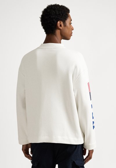 Sweatshirt branca, oversized, com gola redonda. Apresenta texto azul e um gráfico de bandeira no braço esquerdo. Tecido macio e texturizado.