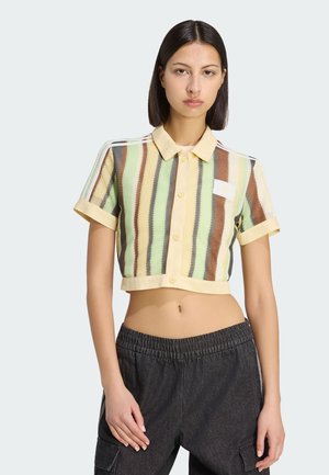 Kobieta ubrana w krótki, zapinany na guziki, krótki top w pionowe zielone, brązowe i beżowe paski oraz czarne spodnie cargo.