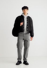 Νέος άνδρας που μεταφέρει μαύρη τσάντα πάνω στον ώμο, φορώντας μαύρο σακάκι, γκρι κουκούλα, γκρι παντελόνι cargo και μαύρα αθλητικά παπούτσια, στέκεται μπροστά σε λευκό φόντο.