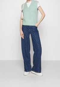 Lichtgroene gebreide mouwloze sweater over een witte t-shirt, gecombineerd met wijde blauwe denimbroek en witte schoenen.