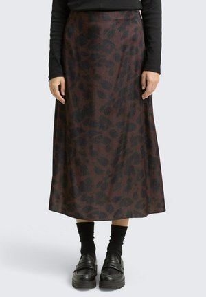 Vrouw in een lange donkerbruine rok met zwarte abstracte vlekken, zwarte sokken, zwarte loafers en een zwart langemouwen top.
