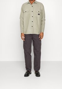 Jasnozielona koszula zapinana na guziki z dwoma kieszeniami na piersi, w połączeniu z ciemnymi spodniami bojówkami z dużymi kieszeniami bocznymi oraz czarnymi butami.