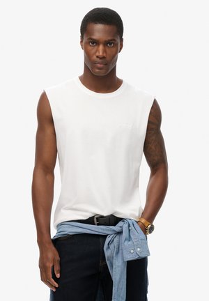 Witte mouwloze T-shirt, gemaakt van katoen, met een ronde hals en een subtiel logo. Gecombineerd met donkere denimjeans en een blauwe blouse om de taille geknoopt.
