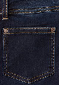 Dunkelblauer Denimstoff mit einer Gesäßtasche, die kontrastierende orangefarbene Nähte und einen Metallnieten in der oberen Ecke aufweist.