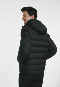 Mężczyzna uśmiechający się, ubrany w czarną kurtkę puchową z kapturem, z rękami w kieszeniach, stojący na tle jednolitego białego tła.