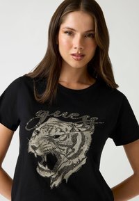 Zwarte t-shirt met een grote, metallic zilveren tijgerafbeelding met rhinestone-accenten en het woord "Guess" erboven in een cursieve lettertype.