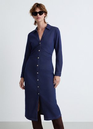 Donna che indossa un vestito camicia blu navy a maniche lunghe con bottoni, occhiali da sole neri e stivali marroni, in piedi su uno sfondo chiaro e uniforme.