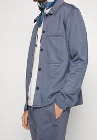 Chaqueta azul con botones frontales, tela texturizada y un solo bolsillo al frente, combinada con pantalones a juego. Presenta botones negros y una marca sutil.