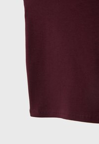 Burgundská bavlněná tričko, krátké rukávy, hladká texturie, rovný lem s detailem šití. Detailní záběr na spodní okraj.