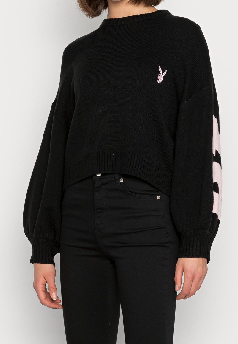 Osoba ubrana w czarny krótki sweter z różowym logo królika na piersi oraz czarne spodnie z wysokim stanem, stojąca na białym tle.