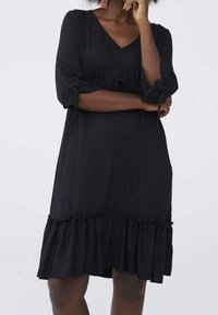 Svart klänning i mjukt tyg med V-ringning och trekvartslånga ärmar. Har samlade detaljer och en volangkant.