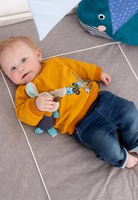 Bambino in un felpa gialla a coste con un design sportivo, che tiene in mano un giocattolo morbido. Jeans in denim blu. Sdraiato su una superficie striata grigia.