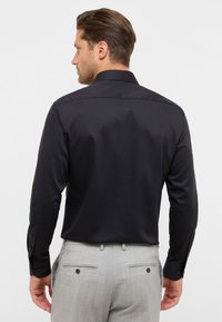 Camicia nera a maniche lunghe con una texture liscia e colletto classico. Con bottoni sui polsini e una vestibilità su misura, abbinata a pantaloni grigio chiaro.