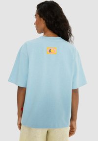 Světle modré bavlněné tričko, volný střih, s barevným grafickým potiskem na zadní straně, krátké rukávy a rovný lem.