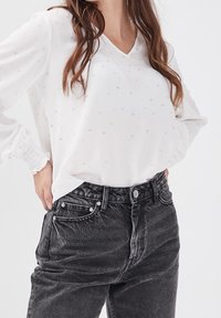 Femme portant une blouse blanche à manches longues avec un motif subtil, rentré dans un jean noir taille haute délavé, les mains dans les poches.