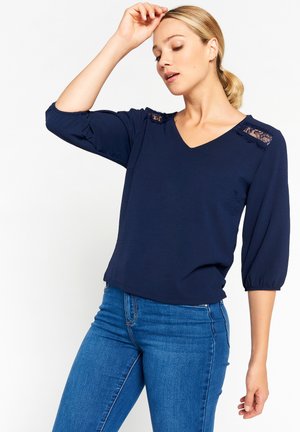 Marineblaue Bluse mit V-Ausschnitt und Dreiviertelärmeln, versehen mit Spitzenakzenten an den Schultern und weichem, strukturiertem Stoff.