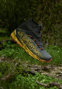 Pantof de alergare pe teren accidentat, de culoare neagră și galbenă, cu crampoane roșii, cu guler mediu la gleznă și marca "La Sportiva", suspendat deasupra ierbii și terenului stâncos.
