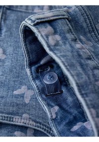 Veste en denim de couleur bleu délavé avec des motifs de papillons, fermeture à boutons et détails de couture visibles. Texture douce et design classique.