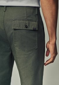 Oliven grønne bomuldsbukser med en baglomme med knaplukning og et lille mærke. Stoffet fremstår glat med minimal tekstur.