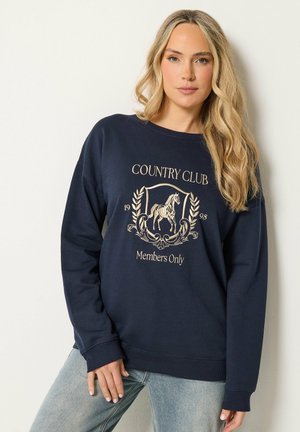 Frau mit langen blonden Haaren, die einen marineblauen Pullover mit einem Pferdeemblem und dem Text "Country Club Members Only 1998" sowie hellblaue Jeans trägt.