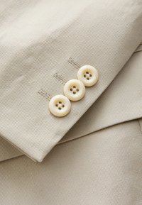 Gros plan de la manche d'une veste en tissu beige avec trois boutons couleur crème alignés verticalement près du poignet.