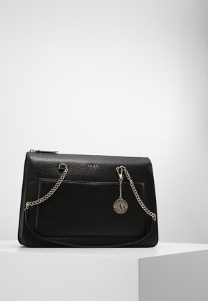 Sac à main DKNY en cuir noir avec bandoulière chaîne argentée, poche avant et pendentif logo circulaire, présenté sur une surface blanche avec un fond gris.