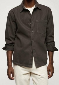 Man in een donkergroene button-up shirt met een borstzak over een wit shirt en off-white broek, met opgerolde mouwen tot aan de ellebogen.