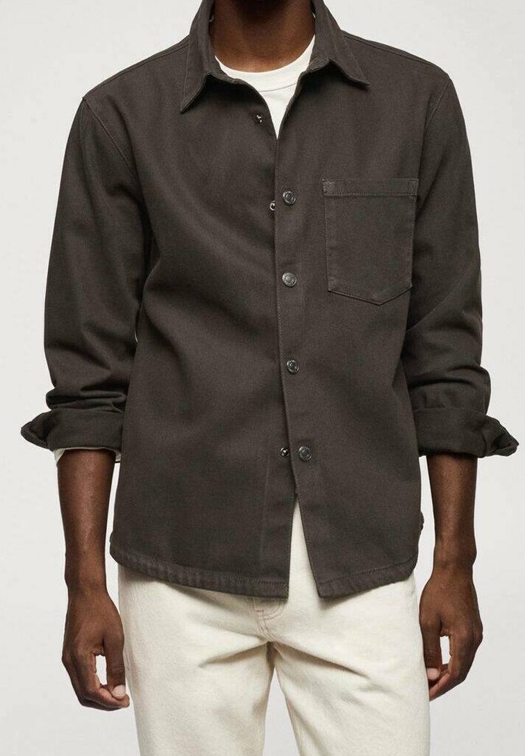 Man in een donkergroene button-up shirt met een borstzak over een wit shirt en off-white broek, met opgerolde mouwen tot aan de ellebogen.
