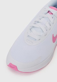 Vit tygidrottssko med texturerad ovandel, rosa detaljer på sulan och snörena, samt en slät tådel med en diskret Nike-logotyp.
