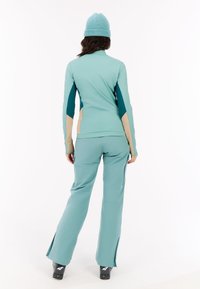 Osoba oblečená v tyrkysovom lyžiarskom oblečení s béžovými a tmavozelenými akcentmi, stojaca so zadočkom otočeným k divákovi, v čiernych lyžiarskych topánkach na bielom pozadí.