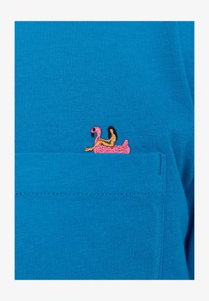 T-shirt bleu avec un design de flottaison en flamant rose brodé sur la poche gauche, avec des accents marron et une texture à côtes fines.
