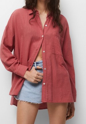 Woman wearing oversized red button-up shirt over light blue frayed denim mini skirt, hand tucked into skirt pocket.