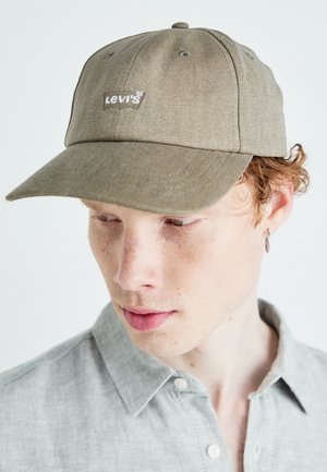 Jeune personne portant une casquette beige Levi's et une chemise à boutons gris clair, regardant vers le bas et sur le côté sur un fond blanc.