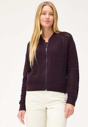 Frau mit langen blonden Haaren, die einen dunkelvioletten gestrickten Reißverschluss-Pullover und weiße Hosen trägt, steht vor einem unifarbenen hellen Hintergrund.