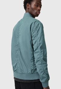 Giacca bomber verde acqua con finitura liscia, polsini e orlo a costine, e dettagli cuciti lungo le maniche e il retro.