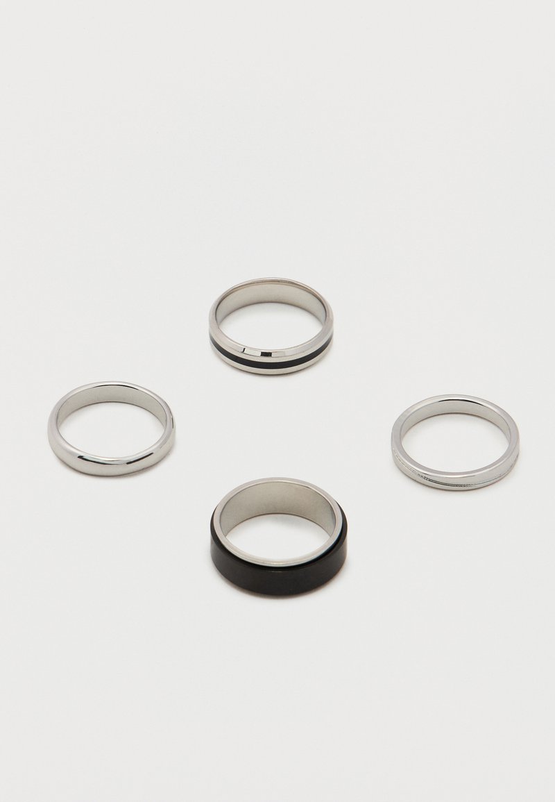 Τέσσερα μεταλλικά δαχτυλίδια σε λευκή επιφάνεια· τρία ασημί με λείες ή ριγέ σχεδιάσεις, ένα φαρδύ με μαύρη εξωτερική ταινία.