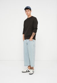 Gebreide zwarte sweater, lichtblauwe wijde jeans en zwart-witte sneakers. Model draagt een denim pet en heeft een zichtbare armtattoo.