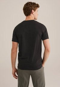 Hombre con cabello rizado castaño, vistiendo una camiseta negra de manga corta y pantalones grises, de pie con una mano en el bolsillo, mirando hacia otro lado.