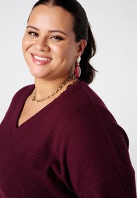 Pull en tricot côtelé bourgogne avec un décolleté en V, doté de bords festonnés. Un collier en chaîne dorée et de grandes boucles d'oreilles rouges en pendentif complètent le look.