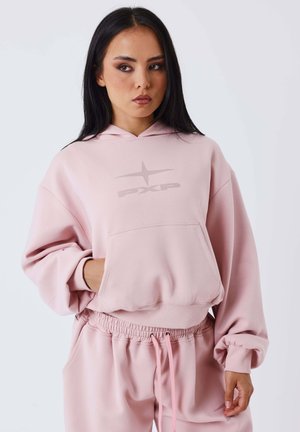 Jeune femme portant un sweat à capuche rose clair et un pantalon de survêtement assorti, avec une main dans la poche du sweat, regardant sur le côté.