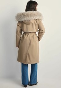 Beżowy płaszcz trench z puszystym, jasnym futrzanym kołnierzem, z paskiem w talii i długimi rękawami, w zestawieniu z niebieskimi dżinsami w stylu dzwonów i czarnymi botkami.