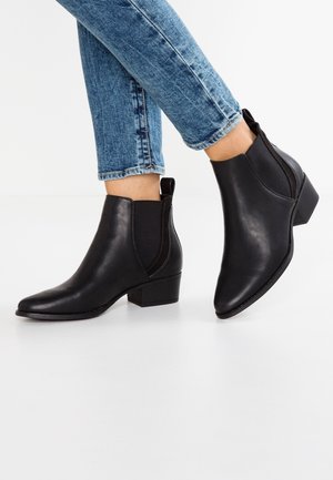 Person iført sorte læderankelstøvler med elastiske sidepaneler og blokhæle, kombineret med blå denimjeans.