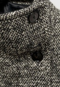 Manteau en laine avec un motif pied-de-poule noir et crème, doté d'un col haut et de grands boutons noirs. Matière texturée et chaude.