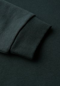 Le tissu du sweat-shirt vert foncé présente un poignet côtelé, harmonieusement intégré à la texture lisse du matériau du corps.