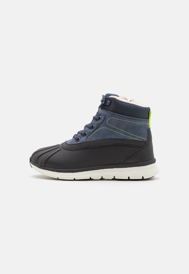 Botte d'hiver montante noire et bleu marine avec semelle blanche, lacets à l'avant et doublure douce en polaire, isolée sur fond gris clair.