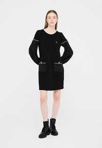 Schwarzes Kleid mit langen, strukturierten Ärmeln und zwei Fronttaschen. Mit goldenen Akzenten und einem runden Halsausschnitt. Kombiniert mit schwarzen Kampfstiefeln.