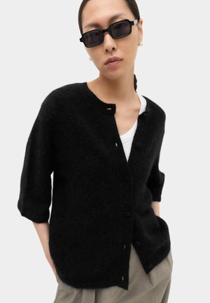 Frau trägt einen schwarzen, flauschigen Cardigan über einem weißen Hemd, beige Hosen und rechteckige schwarze Sonnenbrille, mit zurückgebundenen Haaren.