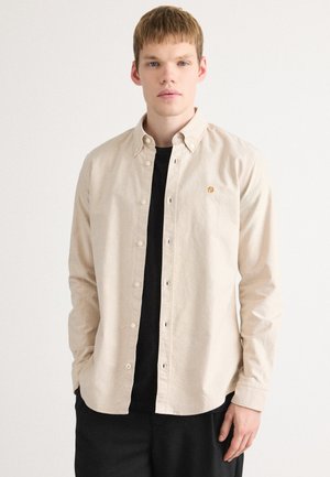 Light beige button-up shirt made of soft fabric, featuring a pointed collar and embroidered logo on the left chest. Worn over a black shirt.